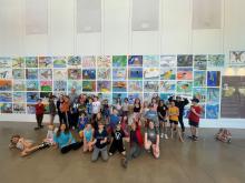 A group of students and one male adult stand in front of a gallery of artwork.