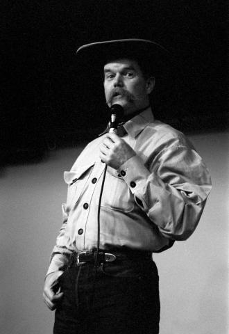 Man with moustache wearing a cowboy hat and white shirt holding a mic on stage. 