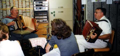 Two older white men seated in a radio station are playing accordions. 