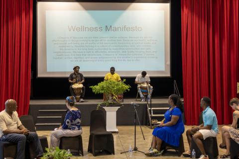 Three people on stage playing drums in front of a large slide projection that say Wellness Manifesto. Others sit in chairs facing the stage.