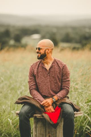 Man with shaved head and beard sitting on a stool smiling. 