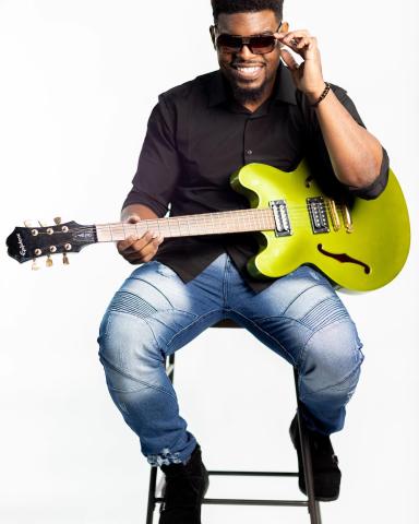 Man wearing sunglasses holding a guitar, sitting. 