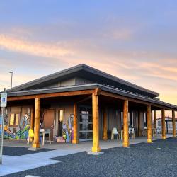 The exterior of a one story building with a colorful mural painted on one side. There are tables and chairs situated under an overhang supported by raw wood columns. The sky is filled with soft pastel pinks, blues, and yellows in what looks to be either sunrise or sunset.