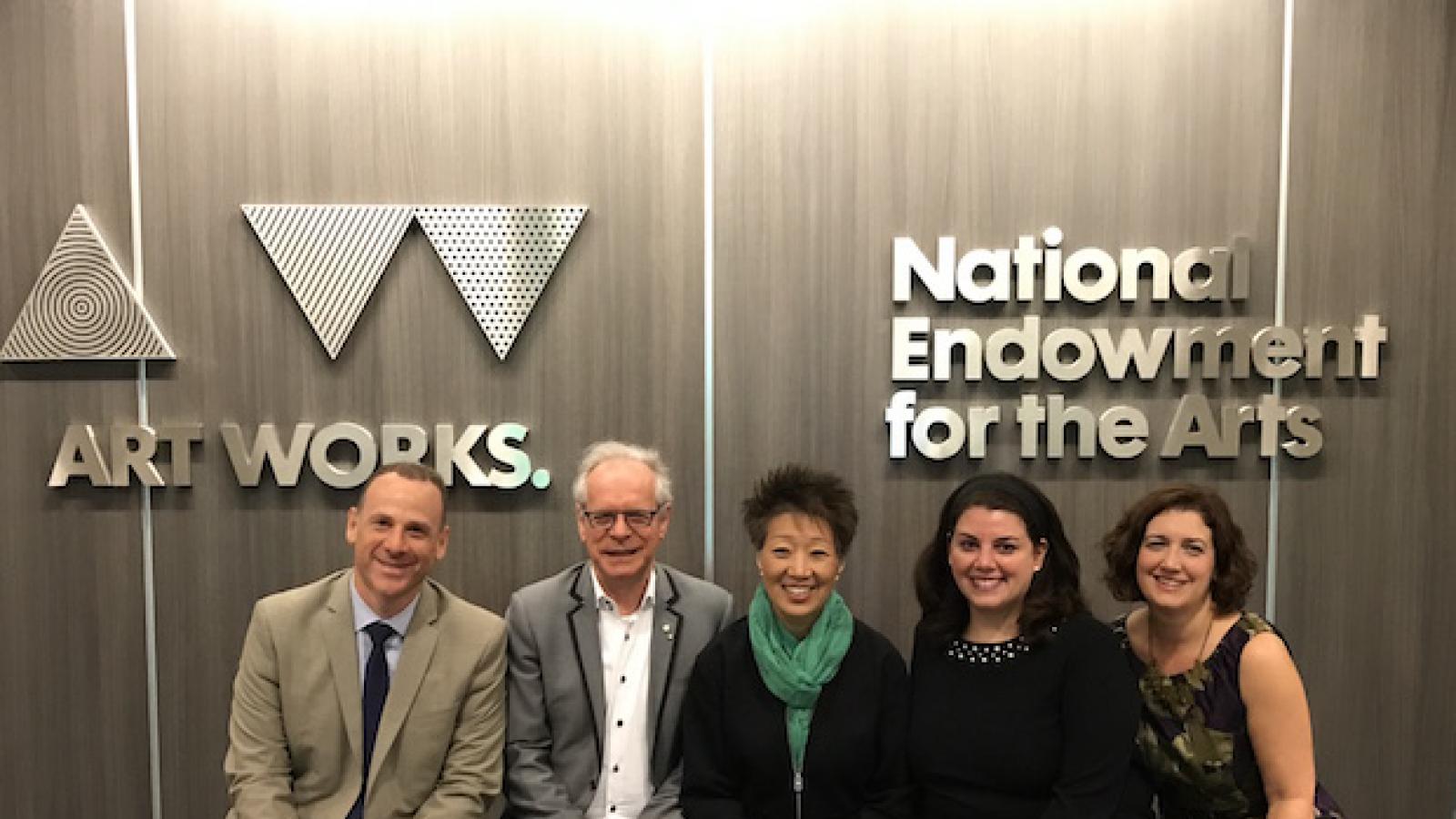 2 white men, an asian woman, and 2 white women  sit on a bench against a background that has the NEA  logo