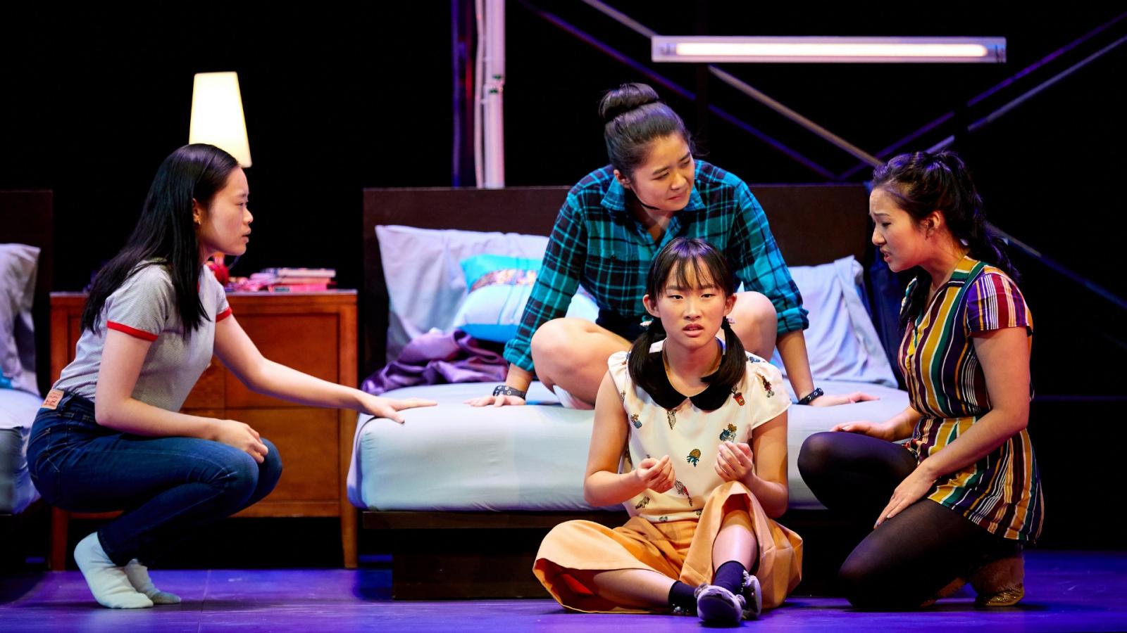 Stage production shot of three Asian women, one on a couch and the other two sqatting, surround an Asian girl. 