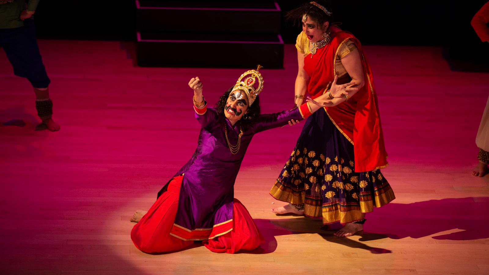 Stage production of two women in colorful clothing, one kneeling wearing fake moustache and shaking fist at sky and the other watching her. 