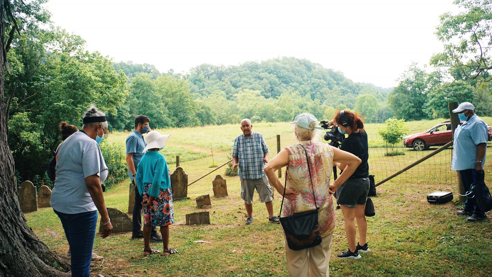 Man in plaid shirt talking to a group of people circled around him in a cemetery. 