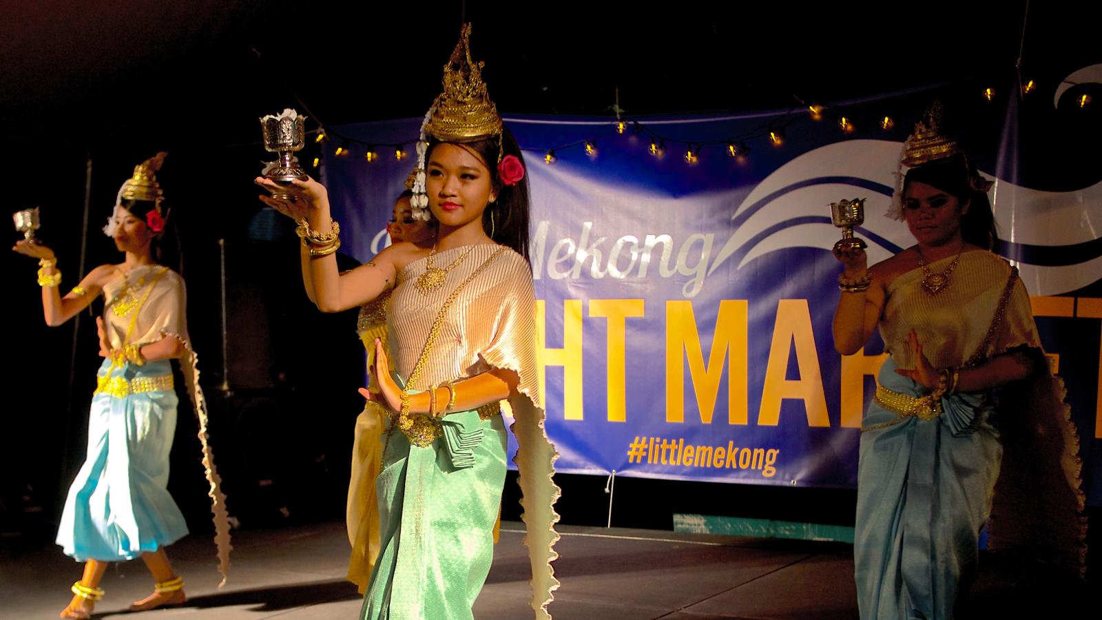 Women in Cambodian garb of blue and yellow in parade down the street, in front of a banner. 