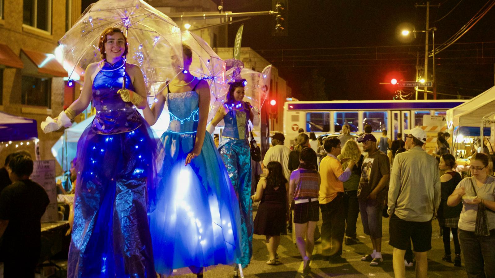Women in blue costumes with lights, carrying umbrellas, walking on stilts through the night market. 
