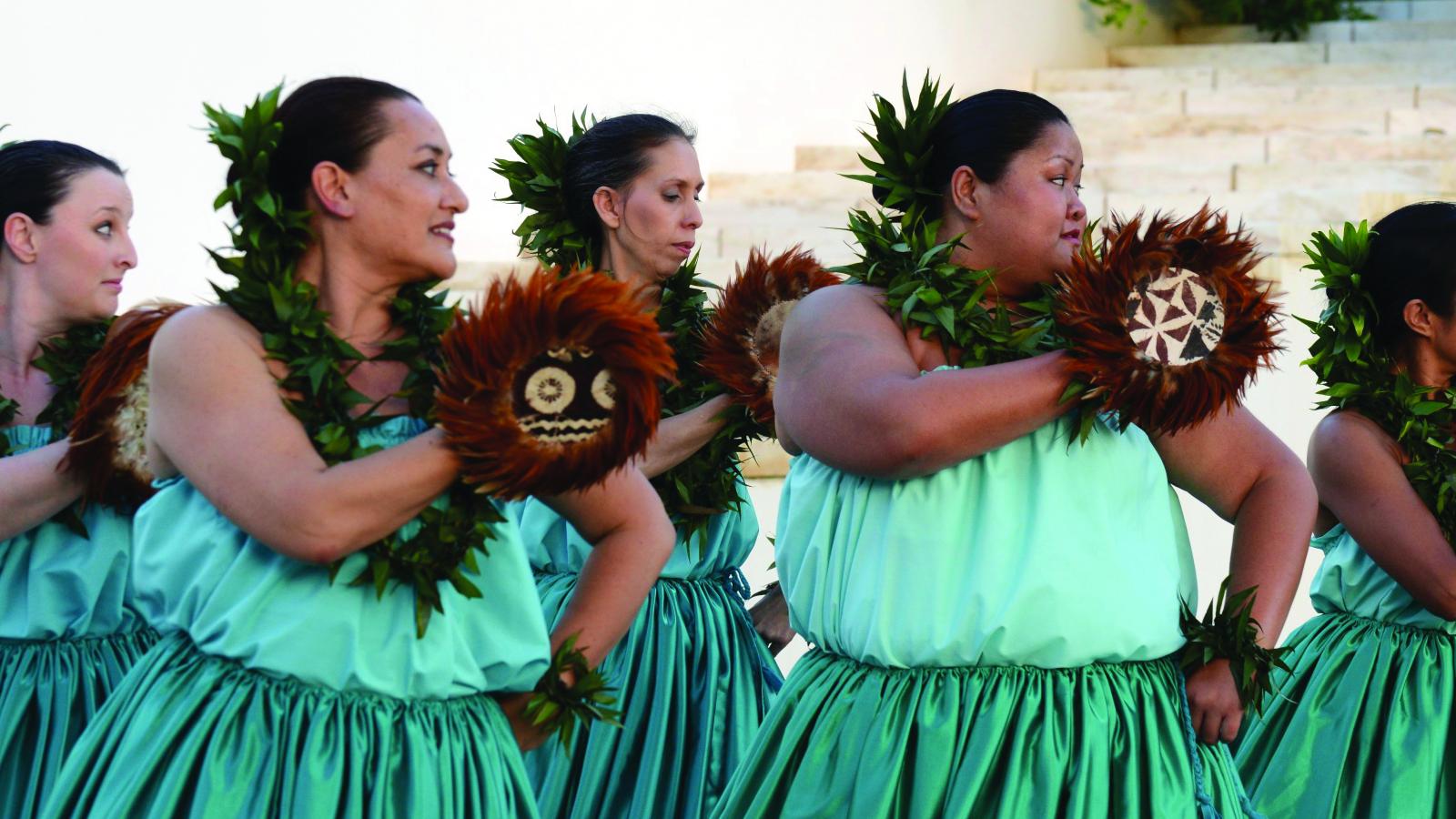Women in blue dresses holding feathered gourds perform a traditional Hawaiian dance.