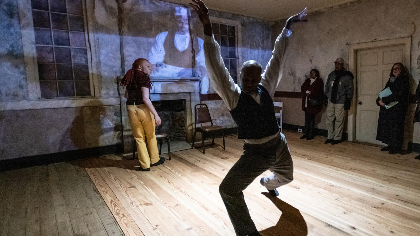 Black man dancing in middle of plantation mansion room with projection on the wall behind him. 