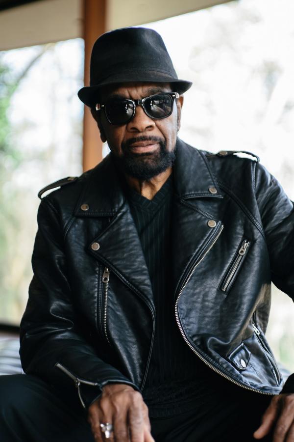Man wearing a pork pie hat, sunglasses, and a leather jacket. 