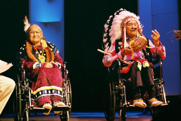 Older Native couple in wheelchairs on stage dressed in traditional Native clothing. 