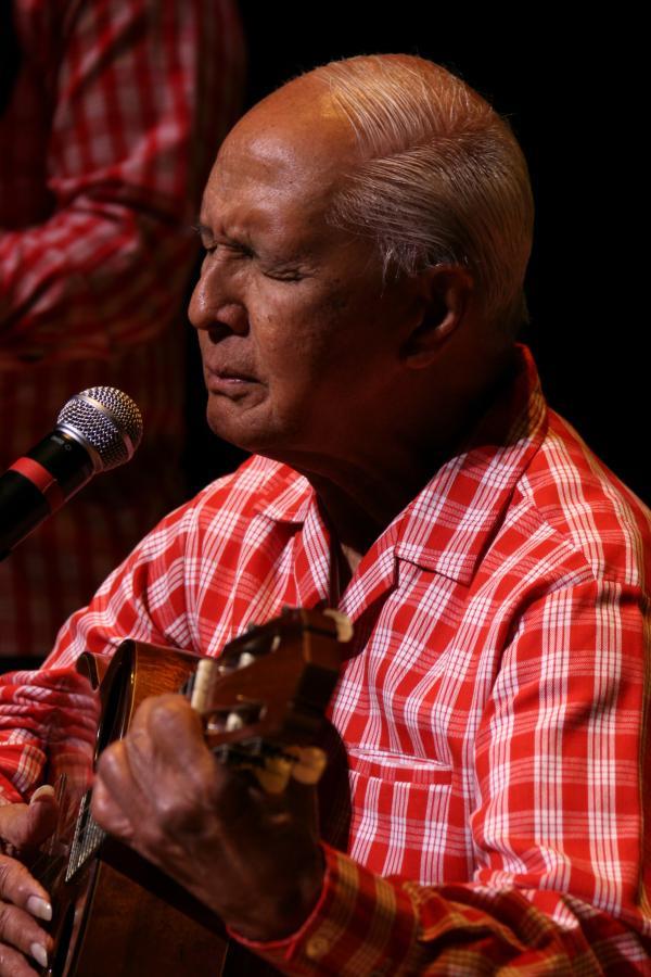 Balding Hawaiian man in red checkered shirt playing a uke. 