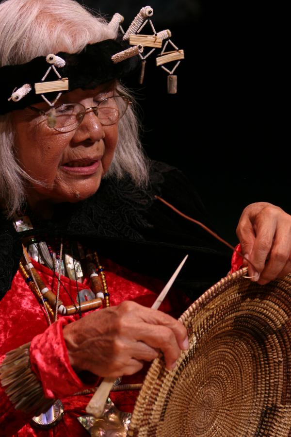 Native woman with white hair wearing headdress and holding up basket. 