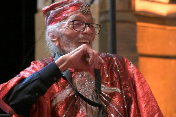 Black man with gray beard and glasses wearing red sequined suit, sitting, laughing. 